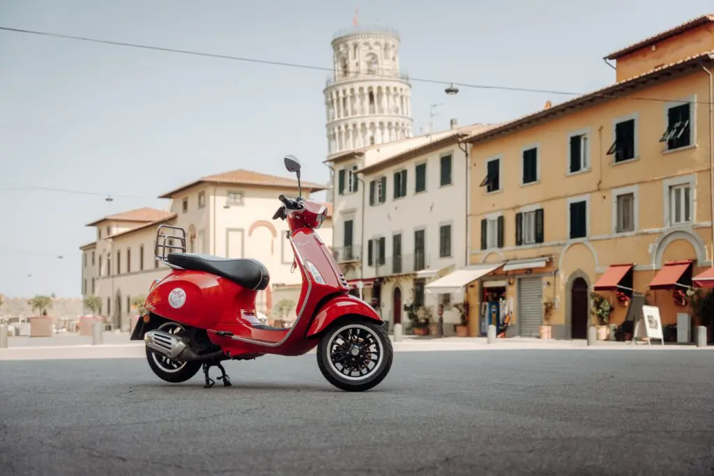 scooter Vespa rouge garé sur une place, avec la tour penchée de Pise