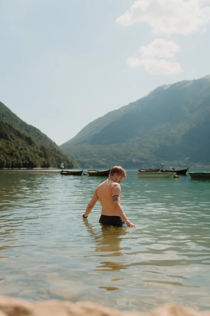 homme avançant dans l’eau d’un lac avec des montagnes et des bateaux