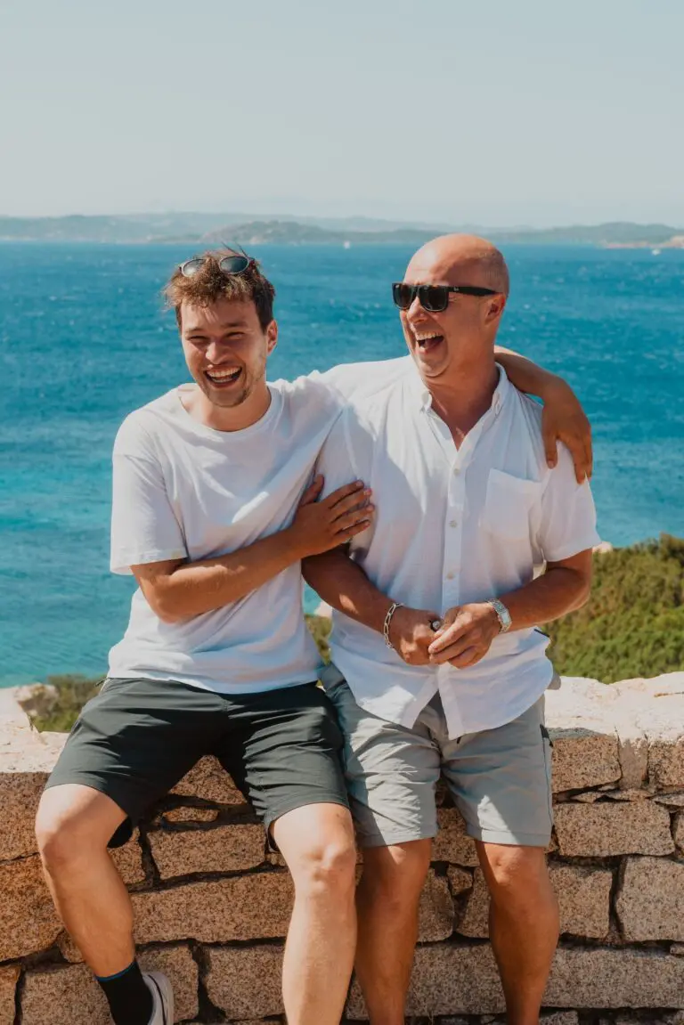 deux hommes rient sur un muret en pierre avec vue sur la mer en Sardaigne